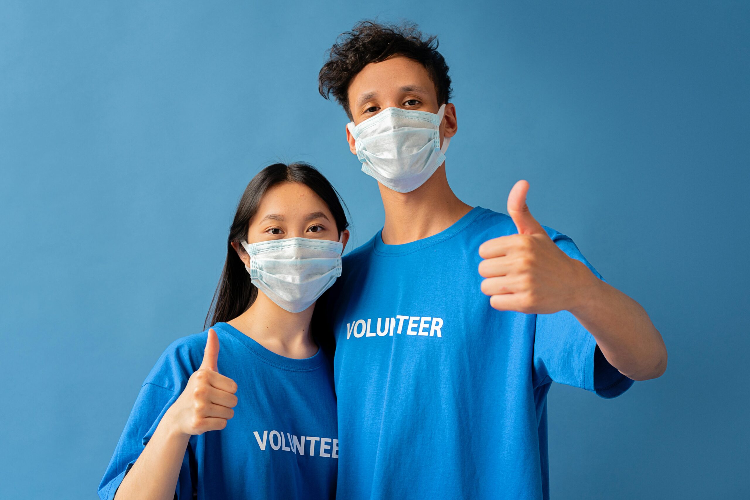 Two volunteers wearing masks and blue shirts giving thumbs up on a blue background.